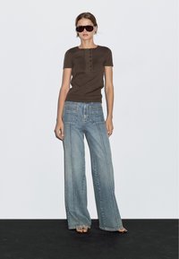 Hnědá přiléhavá košile s krátkým rukávem na knoflíky, spárovaná s volnými světle modrými džíny. Modelka nosí černé sluneční brýle a sandály na pásky.