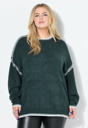 Blonde vrouw in een oversized donkergroene trui met witte bies en zwarte leren broek, staand tegen een witte muur.
