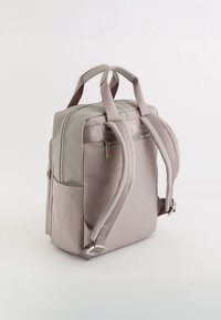 Mochila de tela gris con una parte superior gris suave, dos correas ajustables y acolchadas, un bolsillo frontal con cremallera y un detalle discreto del logo de la marca.