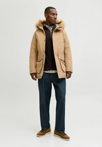 Beige Parka mit einer Kunstpelzkapuze, Reiß- und Druckknopfverschluss, zwei Fronttaschen, über einem dunklen Pullover getragen, kombiniert mit dunkler Jeans und braunen Schuhen.