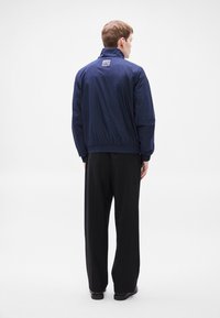 Μπλε σκούρο μπουφάν αδιάβροχο με φερμουάρ, ελαστικά μανσέτες και μέση. Διαθέτει μικρό λογότυπο στη πάνω πλάτη. Φοριέται με μαύρο παντελόνι.