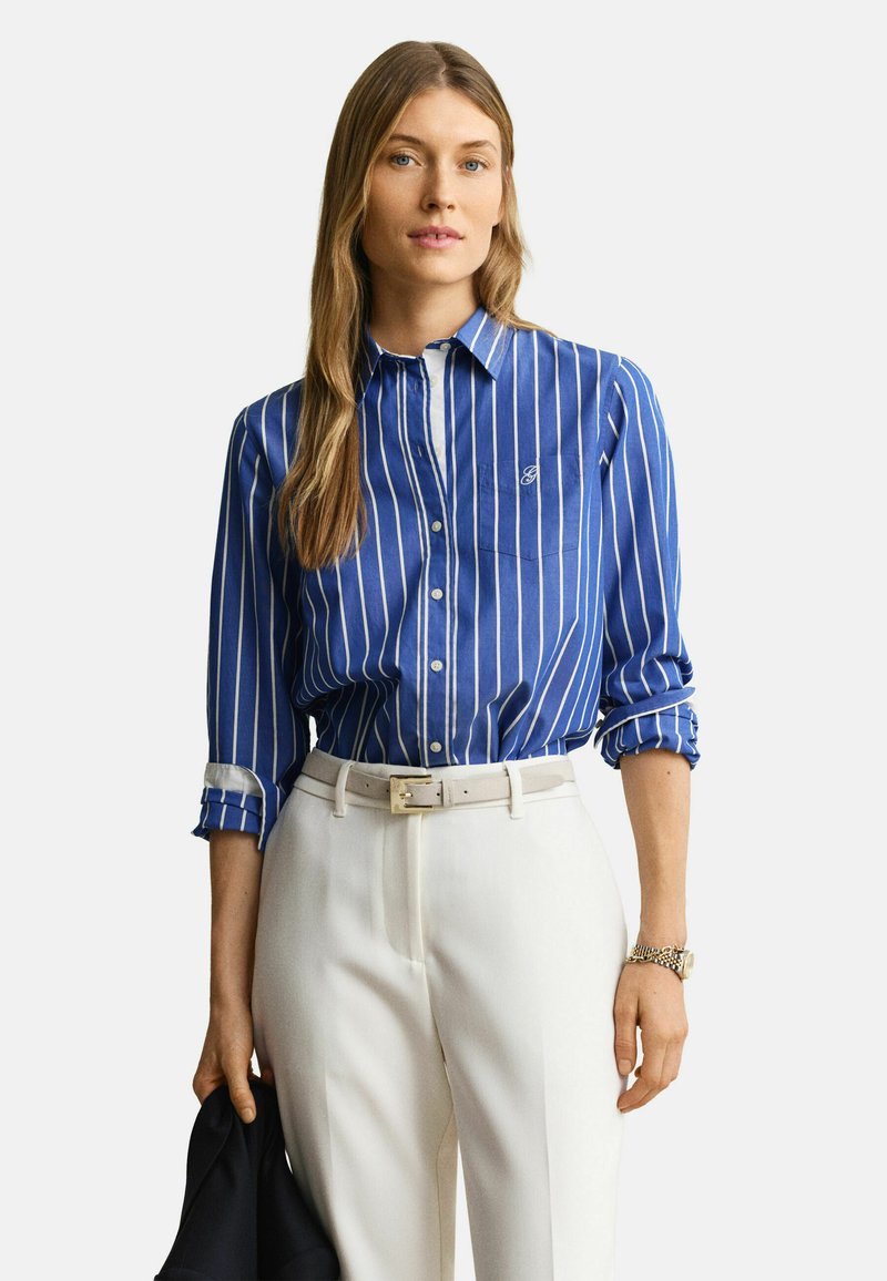 Femme portant une chemise à rayures verticales bleu et blanc, rentrée dans un pantalon blanc taille haute, tenant une veste sombre dans une main.