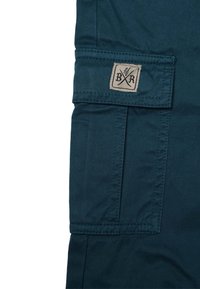 Tumman teal-värinen kangaspokkari, jossa on suorakulmainen läppä ja beige logo, jossa on ristikkäin olevat työkalut sekä kirjaimet "B" ja "R".