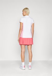 Balta polo marškinėliai su trumpomis rankovėmis, rožinė volanangų sijonas ir rožiniai gėlių akcentai šonuose, derinami su baltais sportiniais batais ir kojinėmis.