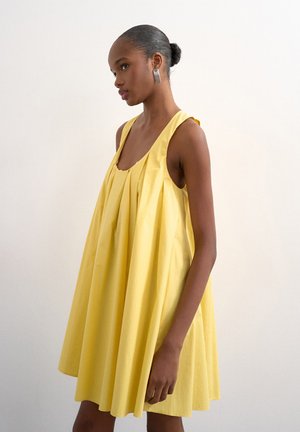 Femme à la peau foncée et aux cheveux relevés en un chignon, portant des boucles d'oreilles rectangulaires argentées et une robe jaune ample sans manches, debout contre un mur blanc uni.
