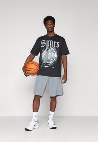 Czarna koszulka z grafiką z napisem "San Antonio Spurs", zestawiona z szarymi szortami. Model trzyma w rękach koszykówkę i ma na sobie białe buty oraz skarpetki Nike.