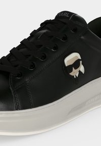 Baskets en cuir noir avec lacets noirs, semelle blanche épaisse, et une petite figurine de dessin animé portant des lunettes de soleil sur le côté.