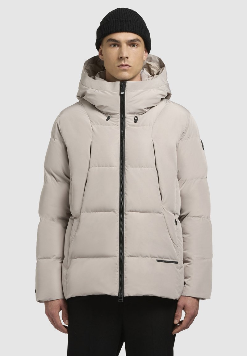 Mann trägt eine hellbeige Daunenjacke mit hohem Kragen und Kapuze, schwarzen Reißverschluss vorne und schwarze Strickmütze vor einem einfarbigen Hintergrund.