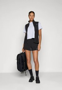 Melns bezroku veste pār gaiši zilu T-kreklu, kopā ar melniem šortiem. Modelis tur melnu mugursomu, valkā tumšas zeķes un sporta apavus.