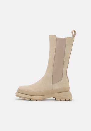 Botte beige mi-mollet avec semelle épaisse, panneau latéral élastique et languette à tirer sur fond gris clair.