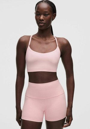 Femme aux cheveux tressés portant un soutien-gorge de sport rose clair et un short de sport taille haute, debout devant un fond uni.