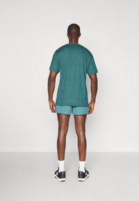 Chemise à manches courtes teal pour homme associée à un short assorti. Le tissu est texturé ; les chaussures sont noires avec des accents blancs. Les chaussettes sont blanches.