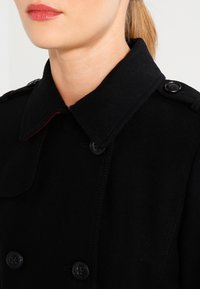 Zwarte wollen jas met een hoge kraag, dubbele knopen, zwarte knopen en contrasterende rode accenten langs de rand van de kraag.