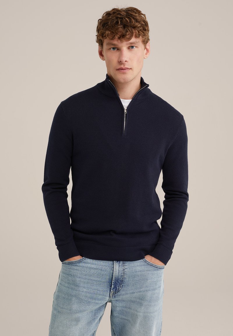 Dunkelblauer Strickpullover mit hohem Kragen und halbseitigem Reißverschluss. Die Textur ist leicht gemustert, mit gerippten Bündchen und Saum.