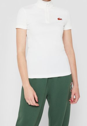 Vrouw draagt een aangesloten wit kort mouwtjes met ritssluiting top met een rood krokodillenlogo en groene broek met meerdere krokodillen patches.