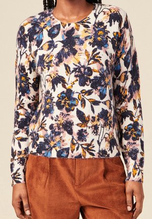 Femme portant un pull à manches longues avec un imprimé floral bleu, orange et noir, associé à un pantalon en velours côtelé marron.