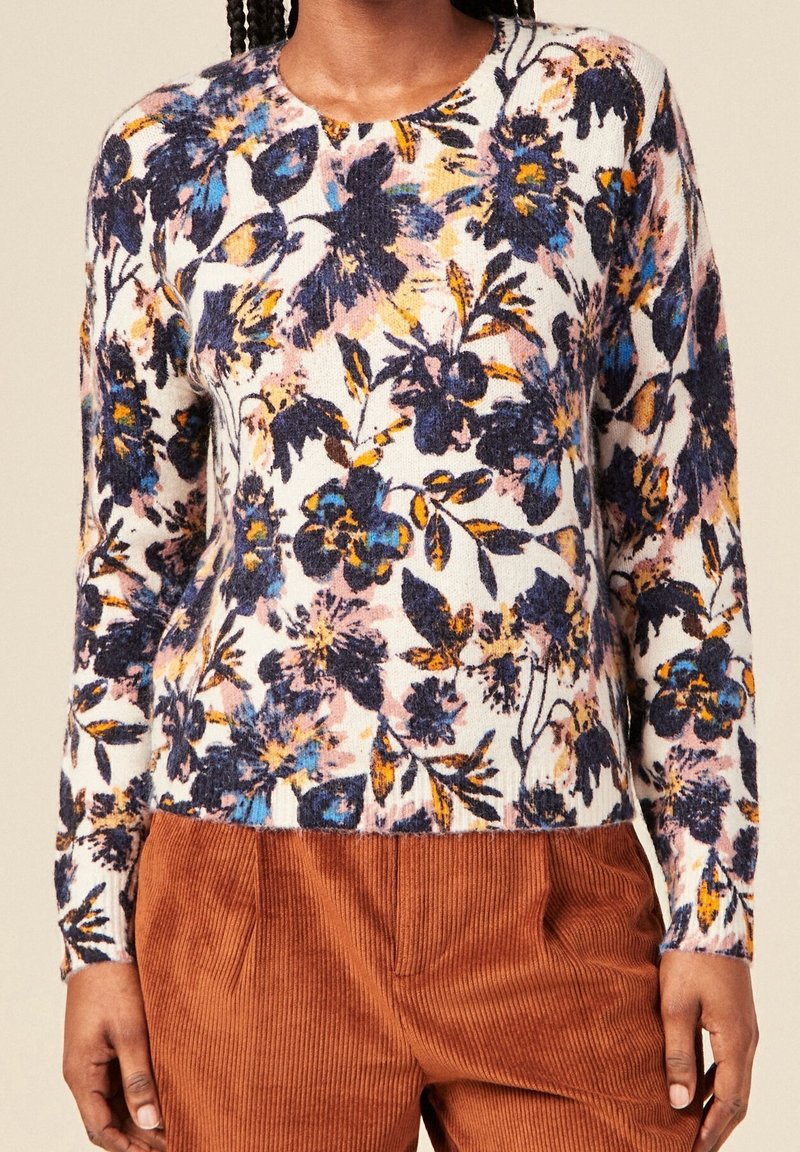 Femme portant un pull à manches longues avec un imprimé floral bleu, orange et noir, associé à un pantalon en velours côtelé marron.