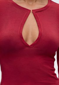 Gros plan d'une personne portant un haut rouge avec un décolleté en forme de trou de serrure montrant le haut de la poitrine et la zone de la clavicule.