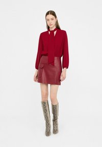 Blusa rossa a maniche lunghe con cravatta al collo, abbinata a una mini gonna in pelle bordeaux a vita alta. Stivali al ginocchio con motivo rettile completano il look.