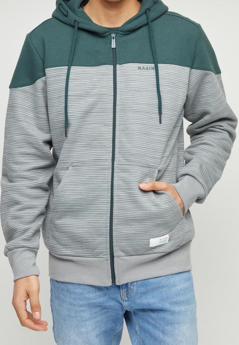 Mężczyzna ubrany w szary i ciemnozielony dżinsowy hoodie z zamkiem, z kapturem i kieszeniami z przodu, w połączeniu z jasnoniebieskimi dżinsami.