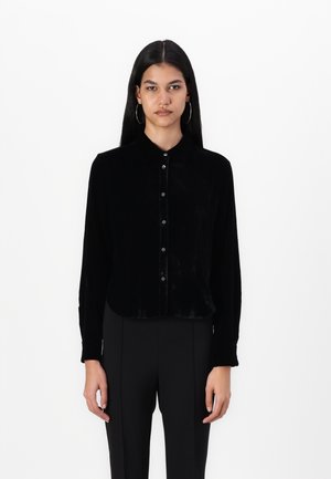 Camisa de veludo preto de mangas longas com frente abotoada, gola e um brilho sutil, combinada com calças pretas ajustadas.