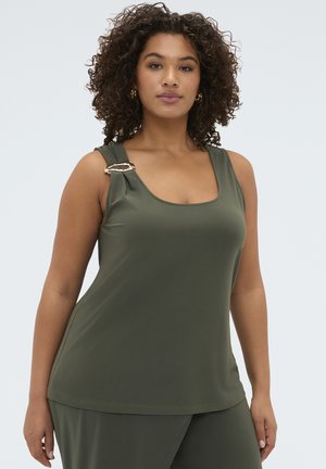 Kobieta z kręconymi włosami, ubrana w oliwkowo-zielony top bez rękawów z złotą klamrą oraz dopasowane spodnie na jednolitym tle.