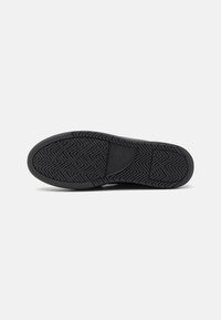 Semelle de chaussure noire avec motif de bande de roulement en zigzag et un patch rectangulaire central avec texte de marque en relief.