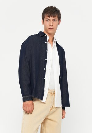 Joven con una camisa de mezclilla azul oscuro sobre una camisa blanca abotonada y pantalones beige claros, de pie frente a un fondo blanco liso.