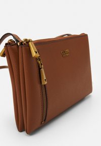 Ruskea текстuroitu nahkak�äsilaukku kultasävyisellä hardwarellä, kaksinkertaisilla vetoketjuilla ja kohokuvioidulla "GUESS EST 1981" -logolla edessä.