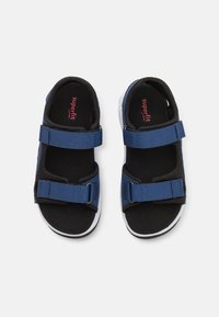 Blå sandaler med två justerbara Velcro-remmar, svarta texturerade innersulor och vita gummisulor. Varumärkesnamnet synligt på innersulorna.