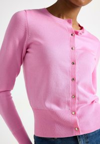 Vaaleanpunainen neuletakki, jossa on pitkät hihat, pyöreä pääntie, hopeinen napitus ja ribbireunus. Valmistettu pehmeästä, kevyestä kankaasta.