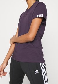 Lila bomullströja med rund halsringning, korta ärmar och vita randar på sidorna av ärmarna, matchad med svarta byxor som har vita randar.