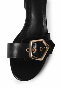Sort lædersandal med en bred strop og en guldfarvet hexagonal spænde, runde udskæringer og en glat tekstur. Minimalistisk design.