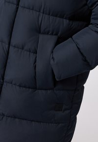 Donkermarine pufferjas met een gequilt design, voorzien van een zijzak en een klein merklogo op de onderste sectie. Glad, glanzend oppervlak.