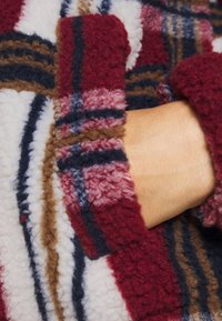 Vêtement en polaire avec un motif à carreaux rouge, blanc, marron et bleu marine. Texture douce avec des poignets retroussés et des détails de couture visibles.