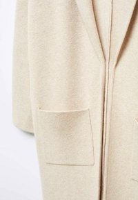 Primer plano de un abrigo de lana beige que muestra dos bolsillos frontales tipo parche y el detalle del cuello con solapa.