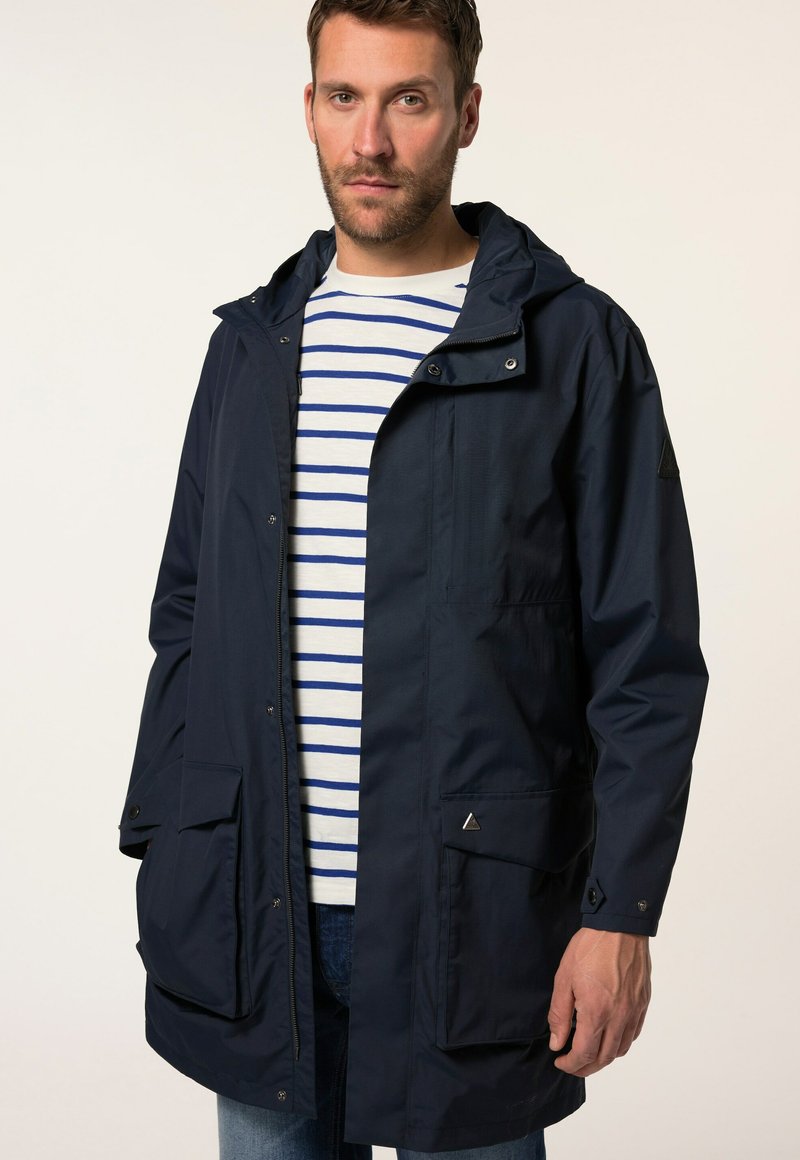 Marineblauwe waterdichte parka met capuchon, ritssluiting aan de voorkant, twee grote klepzakken en een driehoekig accentdetail. Gestreept shirt eronder.