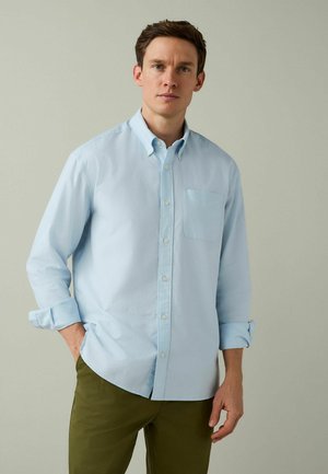 Noor mees helesinises nööbitavas särgis, varrukad üles keeratud, ja oliivrohelised püksid, seistes ühevärvilise halli tausta ees.