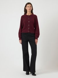 Žena stojaca v burgundskej zapinacej kardigánovej bunde, čiernych širokých nohaviciach, čiernych podpätkových čižmách a zlatých kruhových náušniciach na jednoduchom pozadí.