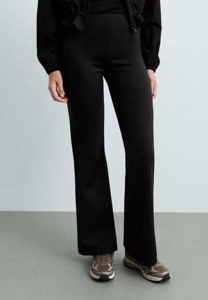 Personne portant un pantalon noir taille haute à jambes larges, une veste noire et des chaussures de sport marron, debout sur un sol gris.