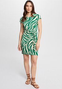 Groene en crèmekleurige gestreepte jurk met een fitted vorm, korte mouwen en een knoopdetail in de taille. G gedragen met bruin gekruiste sandalen.