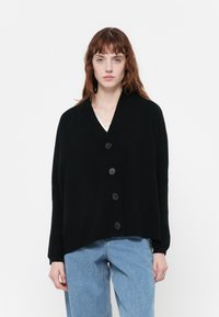 Čierny oversized cardigan vyrobený z mäkkej pleteniny, s výstrihom do V, dlhými rukávmi a piatimi veľkými gombíkmi na prednej strane.