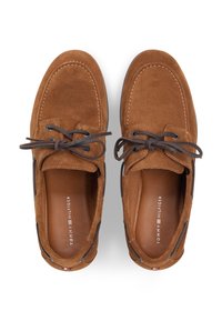 Καφέ σουέτ παπούτσια τύπου boat με σκούρα καφέ κορδόνια, σε αντίθεση ραφές και σχεδιασμό με στρώσεις. Διαθέτουν ενσωματωμένο ταμπελάκι και λογότυπο ως διακοσμητικό στοιχείο.