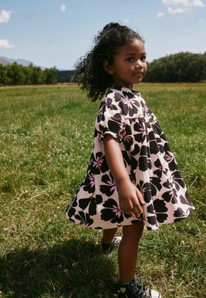 Junges Mädchen mit lockigem Haar trägt ein rosa Kleid mit großen schwarzen Blumen und steht auf einem grasbewachsenen Feld unter klarem Himmel.