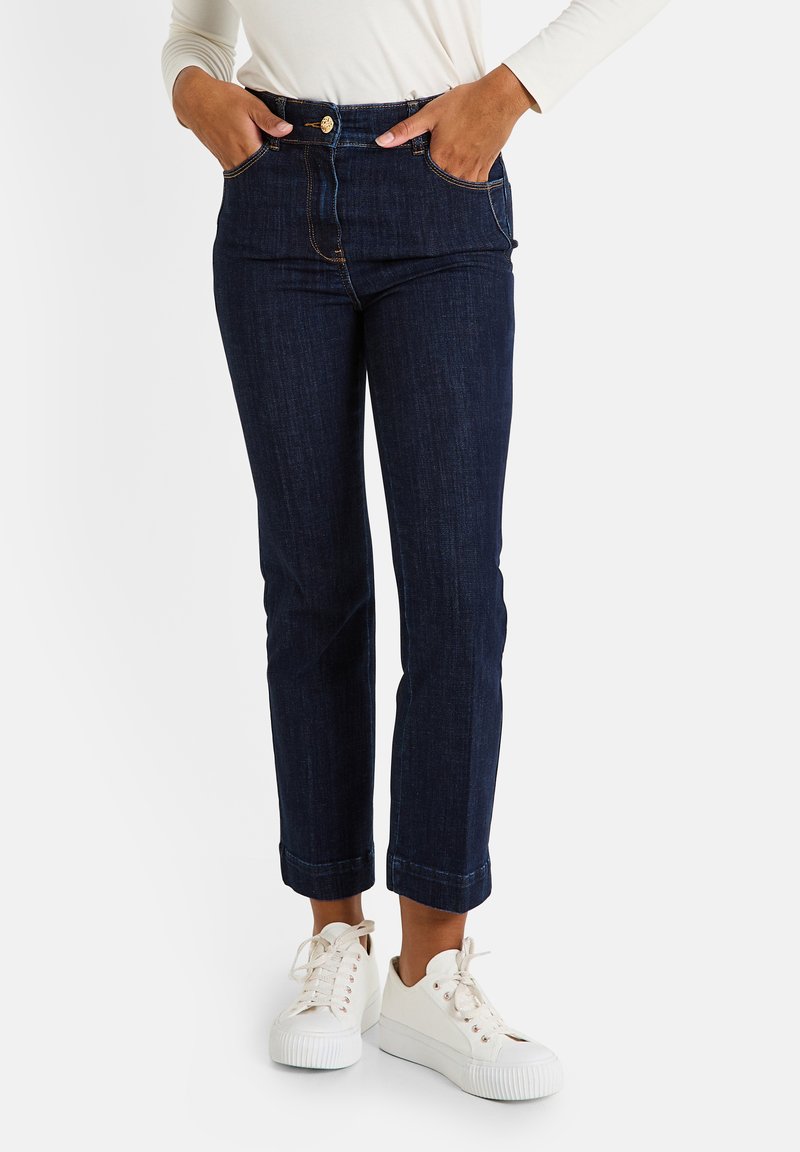 Jean en denim foncé avec une taille ajustée, coupe droite et ourlets roulés. Présente des poches avant et une fermeture à bouton doré. Portés avec des baskets blanches.