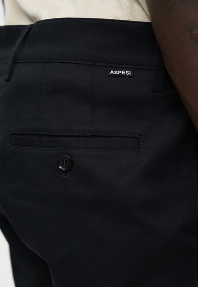 Gros plan sur un pantalon noir avec une poche arrière boutonnée et une petite étiquette de la marque Aspesi près de la taille.