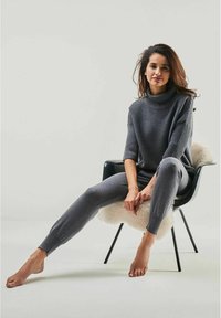 Grauer Strickpullover mit gerippter Textur und hohem Kragen, kombiniert mit passenden grauen Leggings. Model sitzt barfuß auf einem Stuhl.