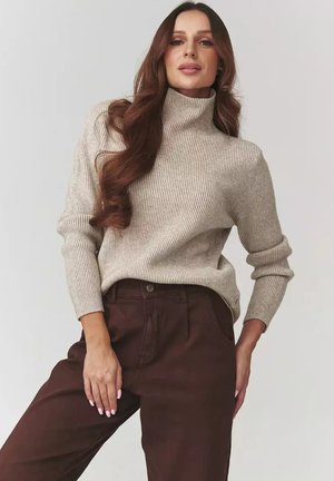 Kobieta z długimi brązowymi włosami ubrana w beżowy sweter z golfem w prążki i ciemne brązowe spodnie na jednolitym tle.