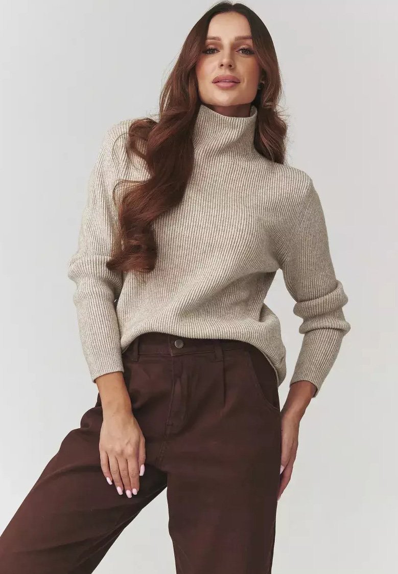 Kobieta z długimi brązowymi włosami ubrana w beżowy sweter z golfem w prążki i ciemne brązowe spodnie na jednolitym tle.