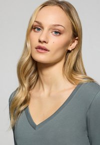 Grå v-ringad topp känd för sin mjuka, släta textur och fitted design. Modellen har blont hår och blå ögon, med diskret smink och lätta örhängen.